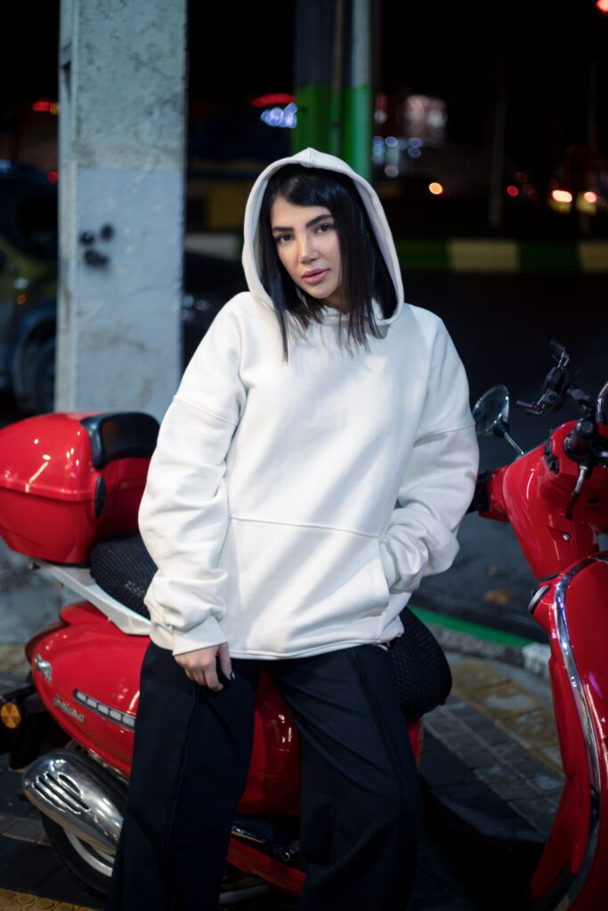 Stylish woman in a white hoodie leans on a red scooter under city lights.