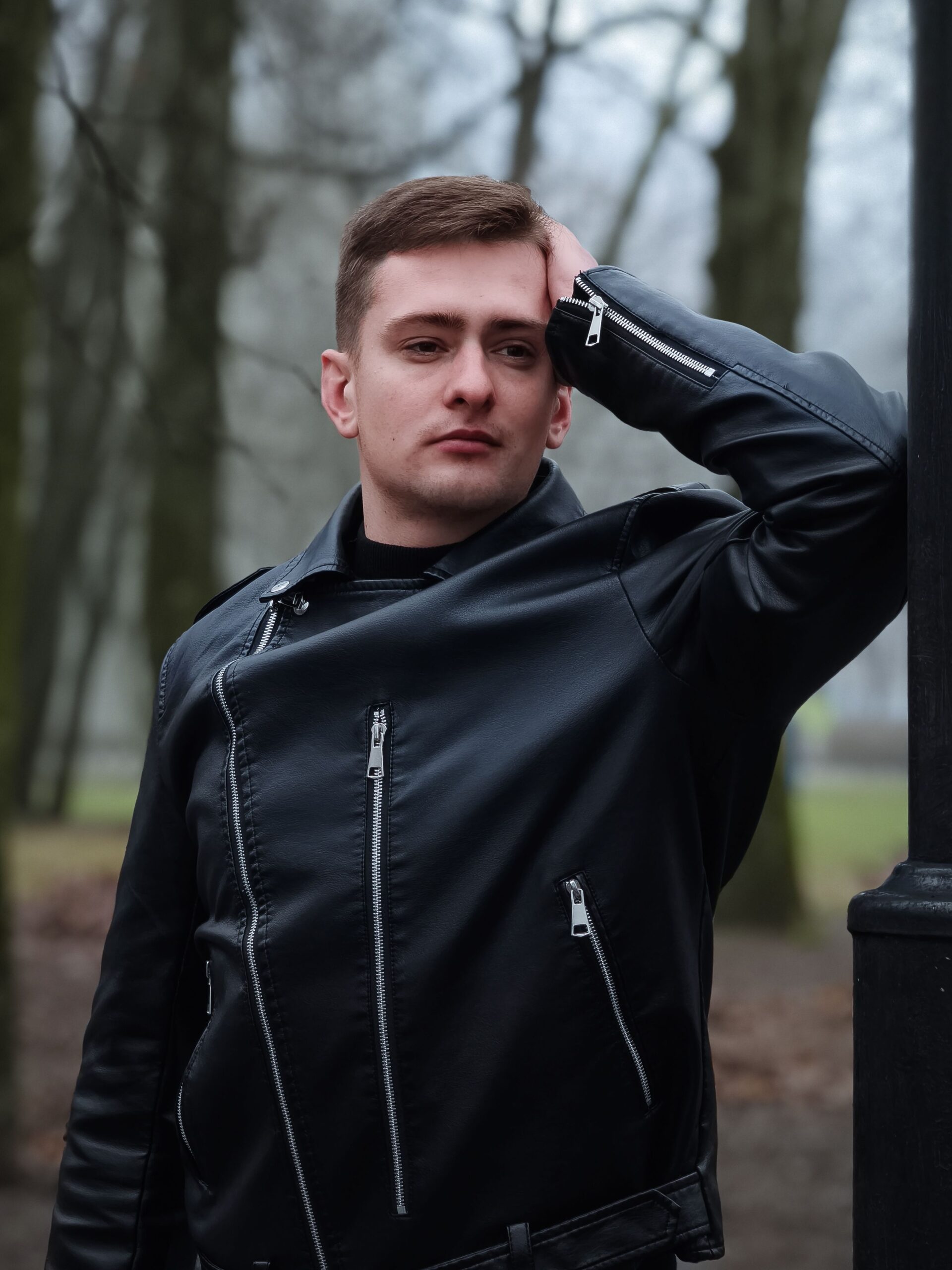 Young man in a leather jacket posing by a lamppost in an autumn park setting.