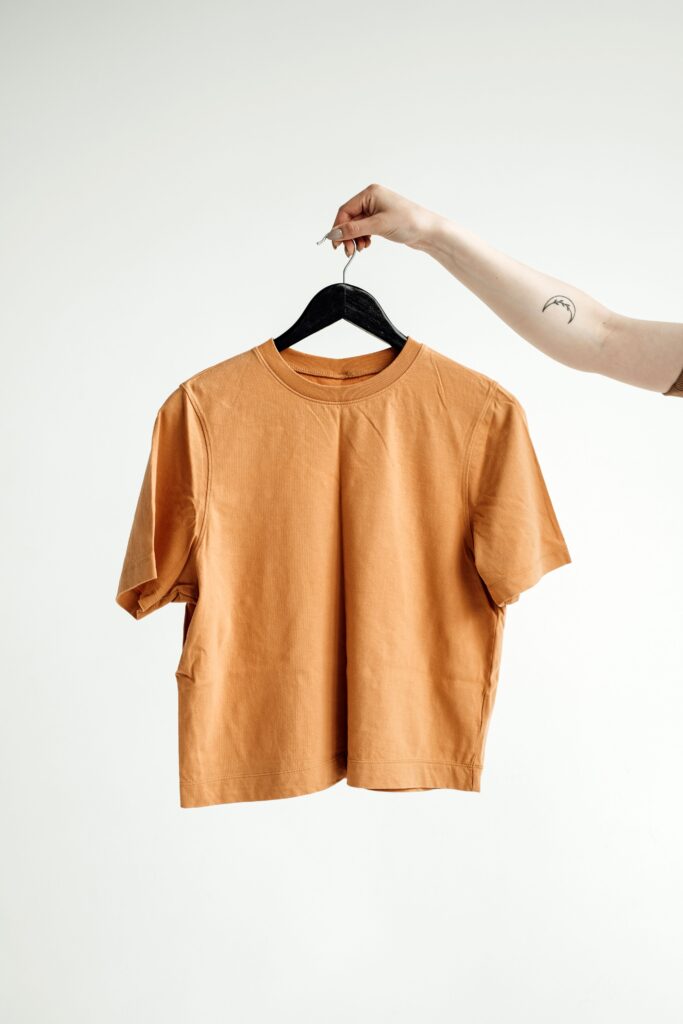 Minimalist shot of an orange t-shirt on a hanger held by a tattooed hand against a white background.